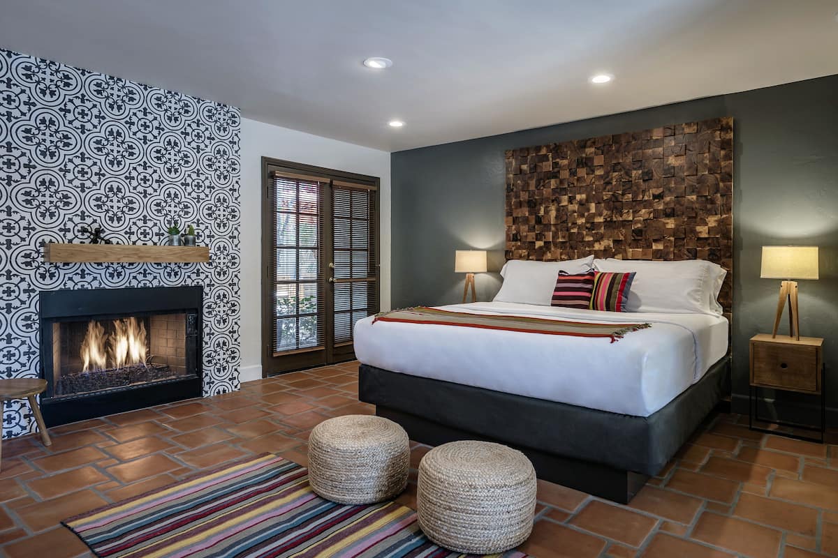 A bedroom with a fireplace, a bed with pillows, two ottomans, a striped rug, and a wall with a geometric pattern.