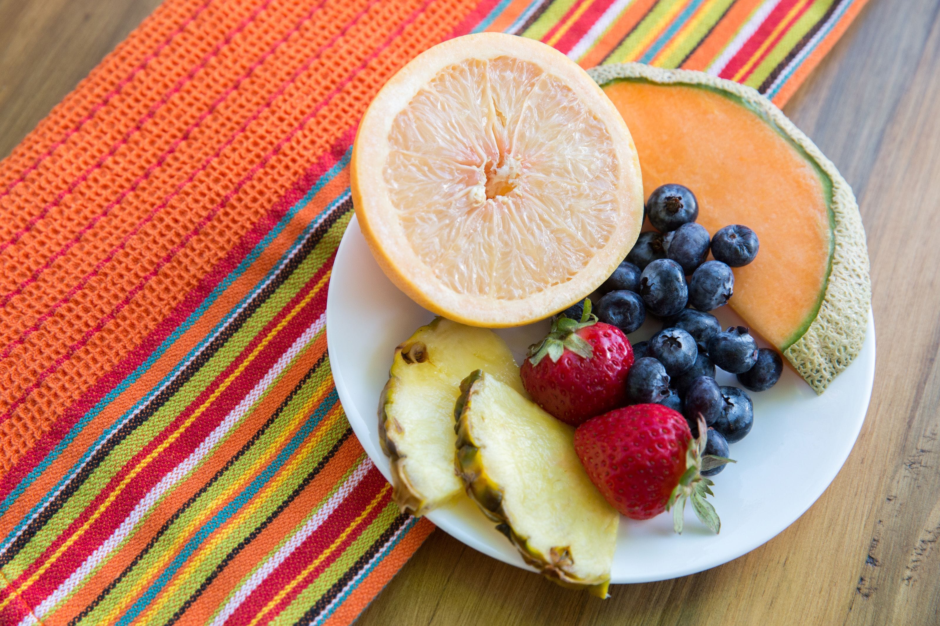 Un piatto con ananas a fette, fragole, mirtilli e un cantalupo tagliato a metà su un telo colorato a righe.