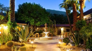 A well-lit garden with a central fountain, surrounded by palm trees and cacti, leading to a building with a red-tiled roof.