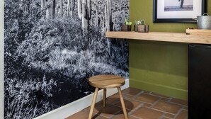 A room with a large black and white cactus mural, a wooden stool, a framed picture on the wall, and a green wall.