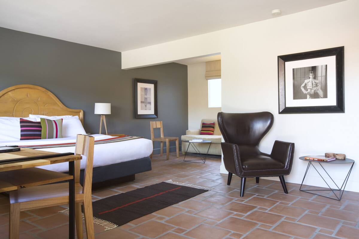 A hotel room with a bed, a desk, a chair, a small table, and a framed picture on the wall.