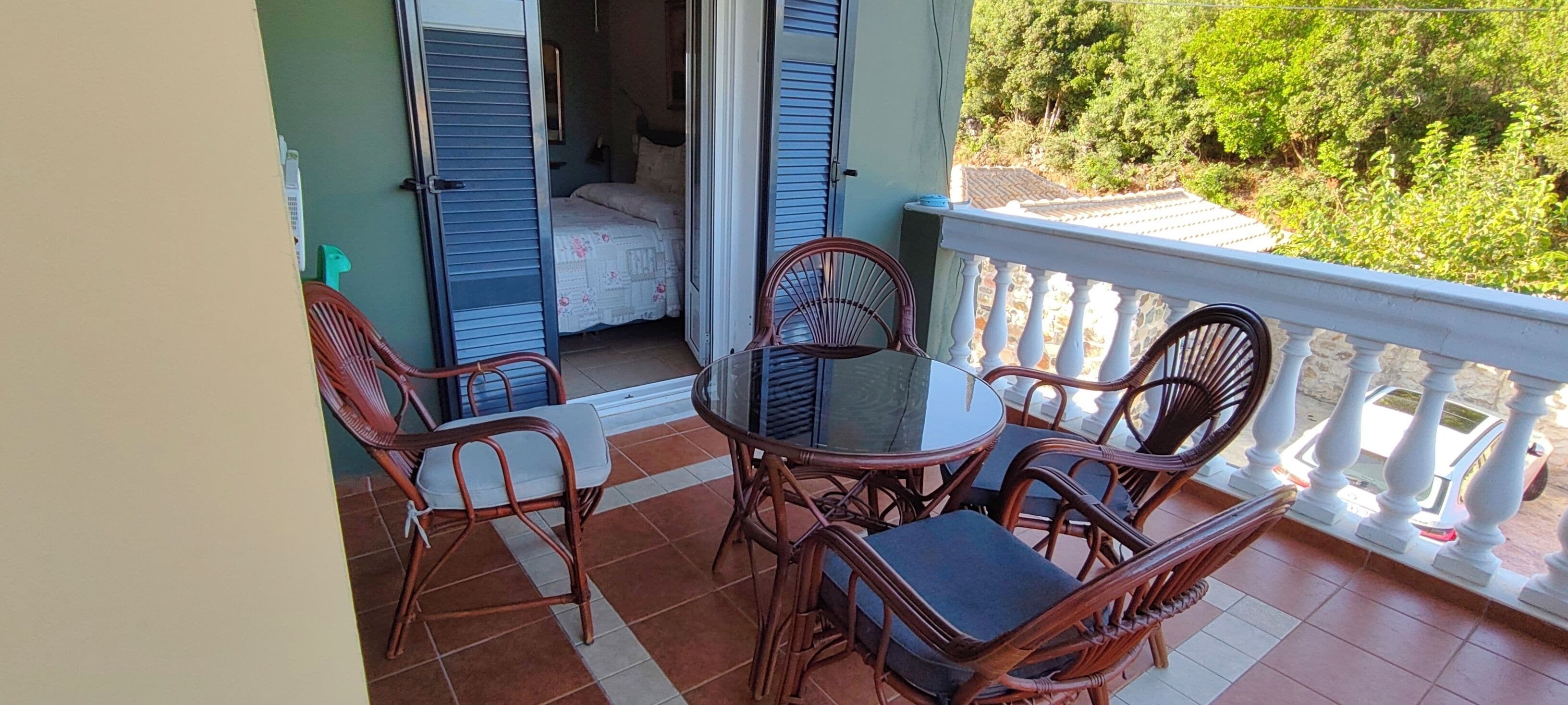 Ein Balkon mit Korbstühlen und einem Glastisch, mit Blick auf einen Garten und ein Gebäude mit blauen Fensterläden.