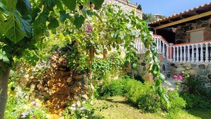 Ein Steinhaus mit weißem Balkon, umgeben von üppigem Grün und blühenden Pflanzen.