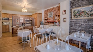 Un comedor acogedor con sillas y mesas blancas, una pared de ladrillo con estantes, y una cocina al fondo.