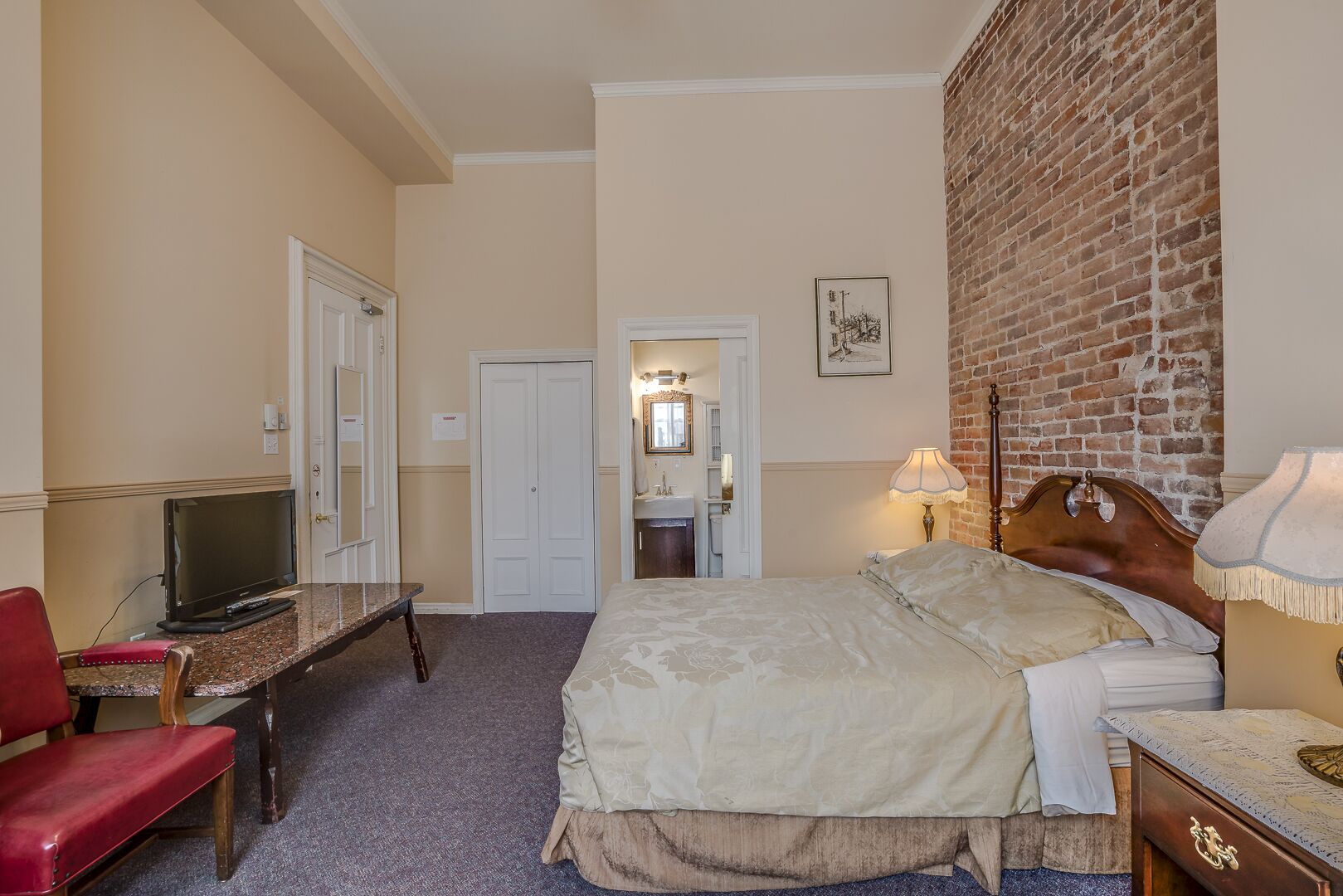 Hotelkamer met een bed, een bureau met een televisie, een rode stoel, een bakstenen muur en een badkamer die door een open deur zichtbaar is.