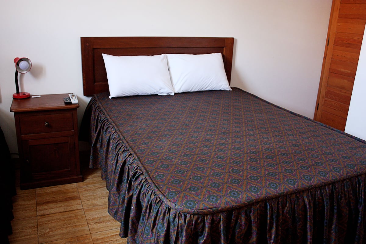 A neatly made bed with a patterned bedspread, a wooden nightstand with a lamp and remote control, and a wooden floor.
