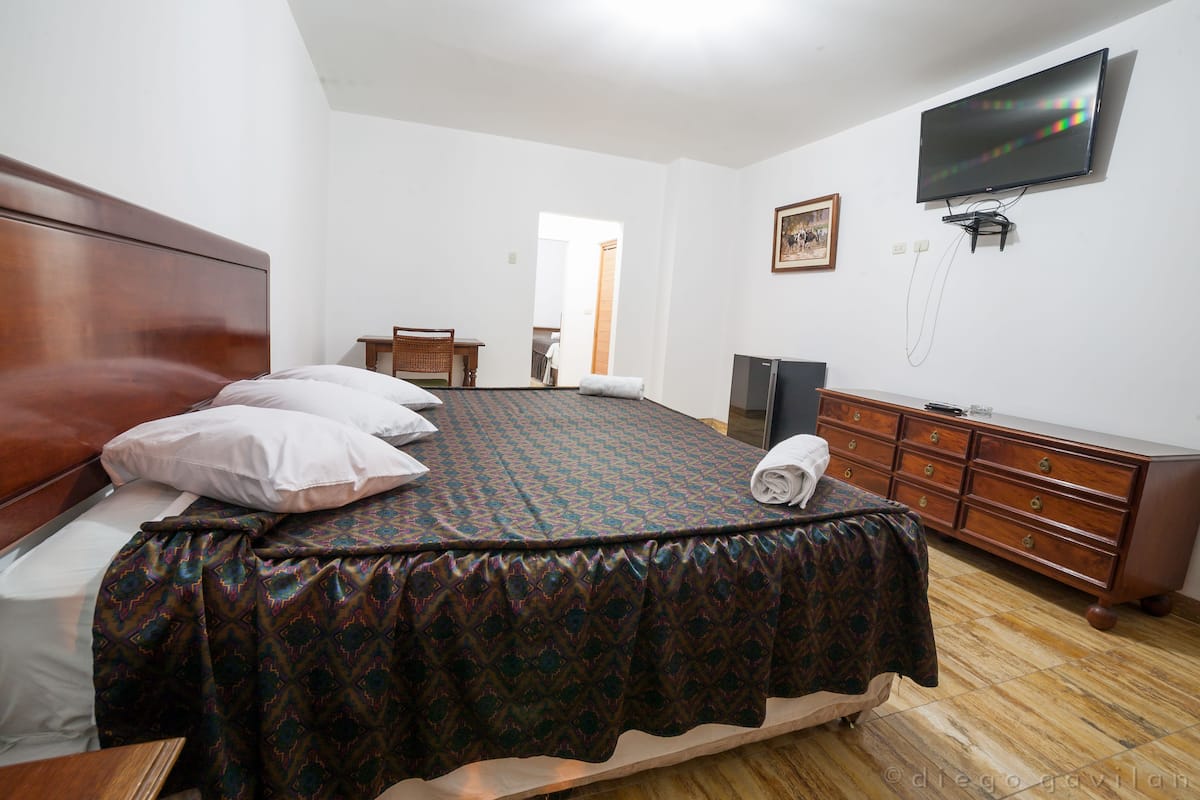 A bedroom with a large bed, a dresser, a television, and a door leading to another room.