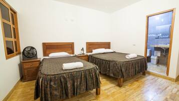 Une chambre d’hôtel avec deux lits, un plancher en bois, un petit ventilateur et une vue sur l’extérieur grâce à une porte vitrée.