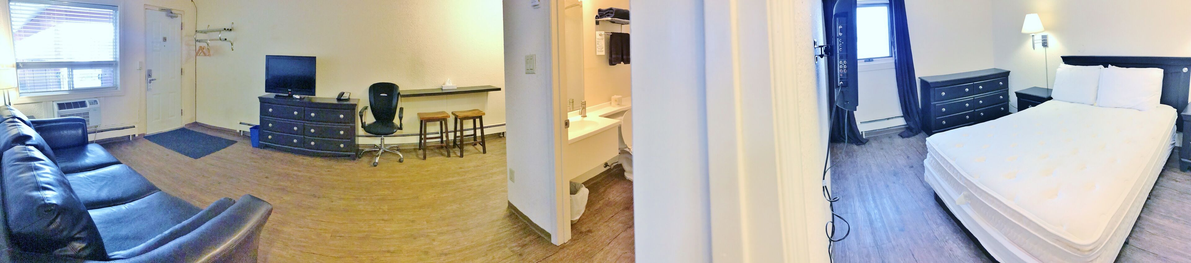 A room with a sofa, a desk with a chair, a small kitchen area, and a bed.