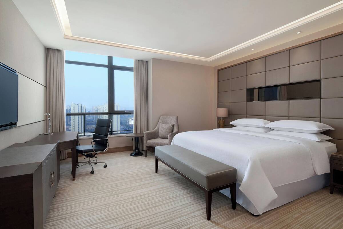 A hotel room with a large bed, a desk, a chair, a bench, a TV, and a view of the city.