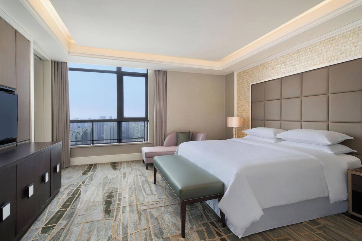 A hotel room with a large bed, a bench, a TV, and a view of the city.