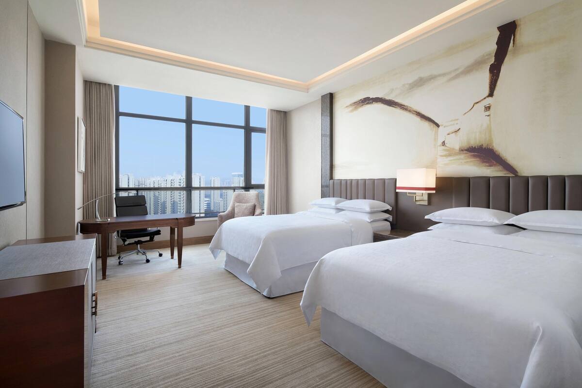 A hotel room with two beds, a desk, a chair, and a large window offering a city view.
