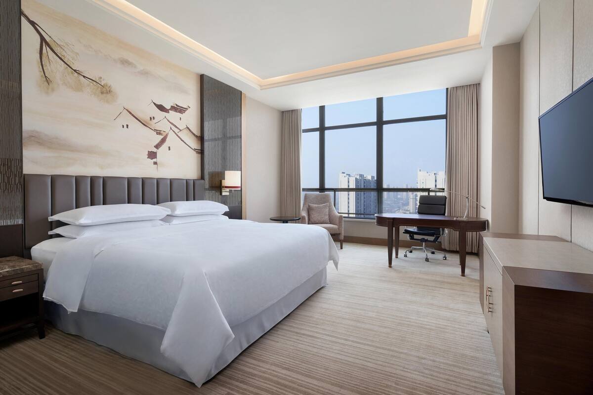 A modern hotel room with a large bed, a desk, a chair, a TV, and a view of the city through large windows.