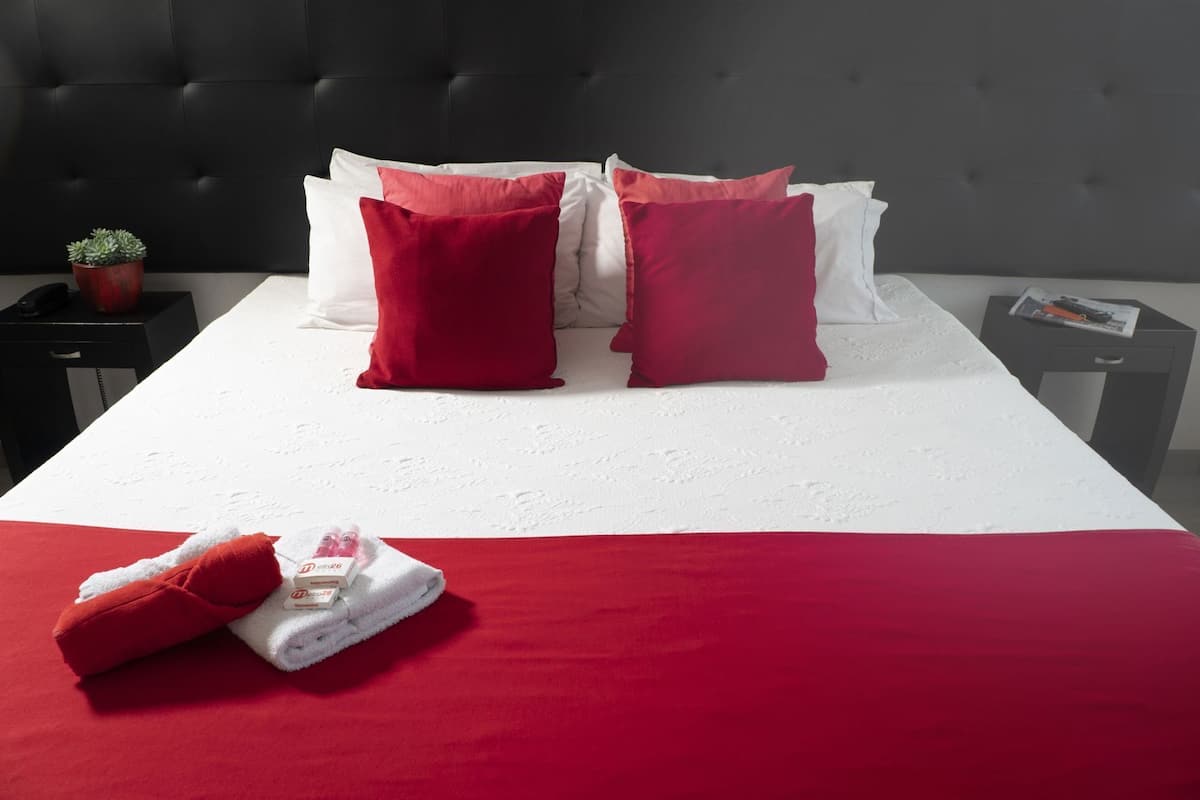 Una cama bien hecha con ropa de cama roja y blanca, una pequeña planta en maceta y una revista en una mesita de noche.