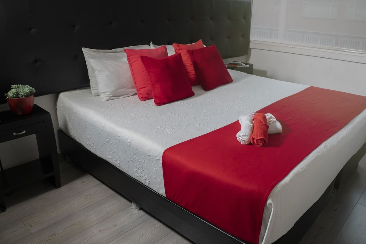 Una cama bien hecha con una colcha blanca y roja, un cabecero gris y una pequeña planta en maceta sobre una mesita de noche.