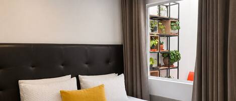 A hotel room with a large bed, a headboard, a bedside table, a window with curtains, and a view of a balcony with plants.