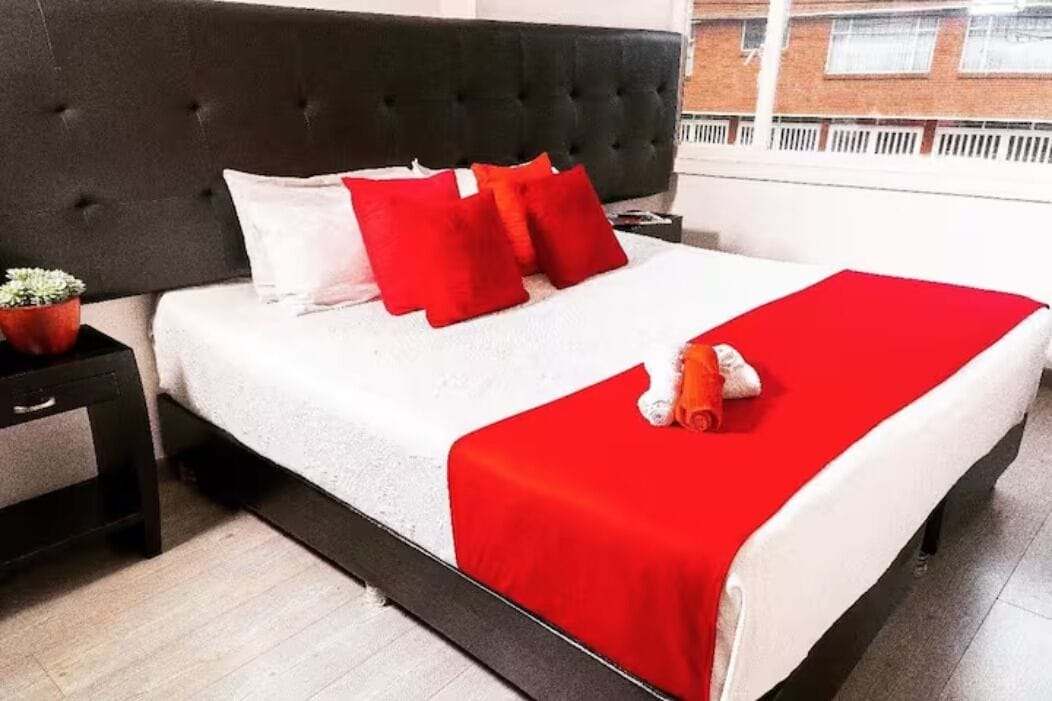 Una cama con cabecera oscura, ropa de cama blanca y detalles en rojo.