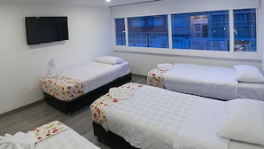 A hotel room with four beds, a flat-screen TV mounted on the wall, and a window with a view of a blue fence and buildings. - Hotel Metro 26 (Bogotá)