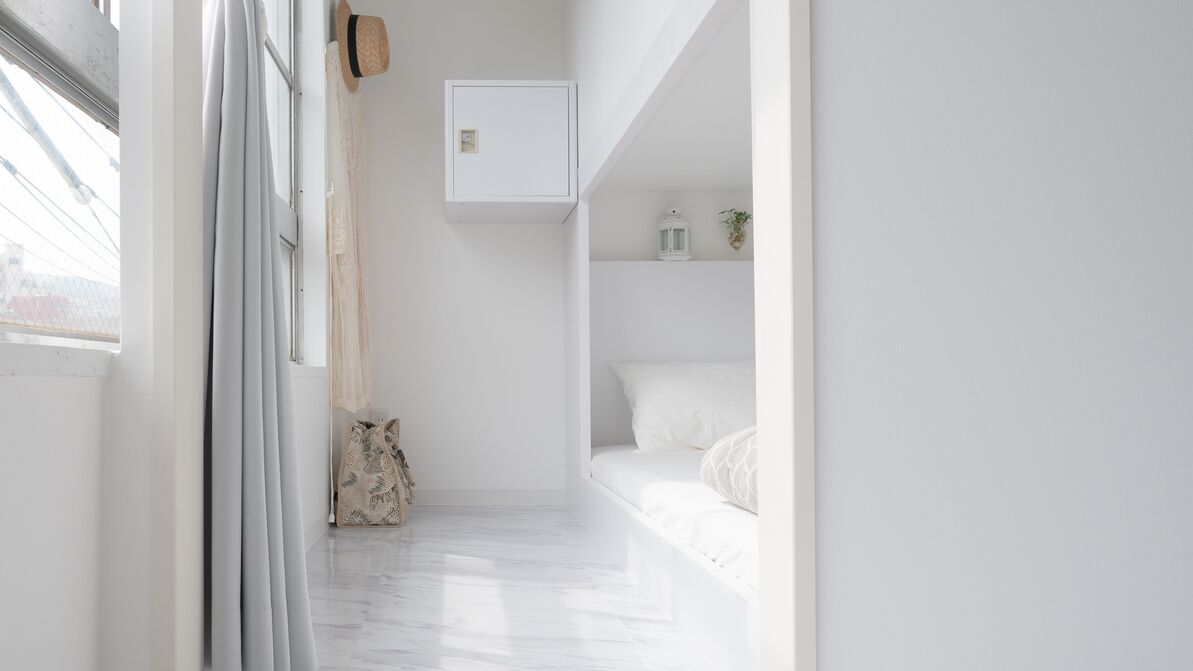 Um corredor estreito leva a um quarto branco com uma cama, um armário pequeno e um chapéu pendurado.