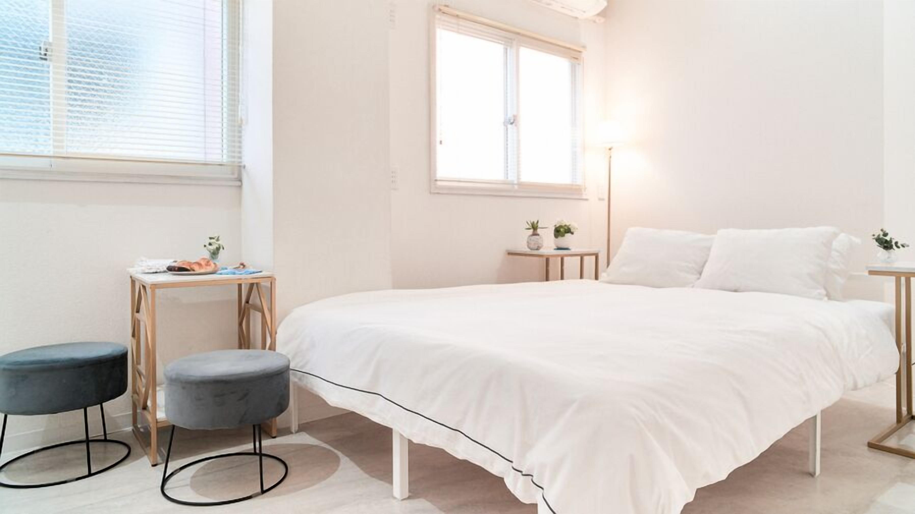 Quarto com uma cama grande, uma escrivaninha, duas cadeiras, uma mesa pequena com uma planta e uma janela com persianas.