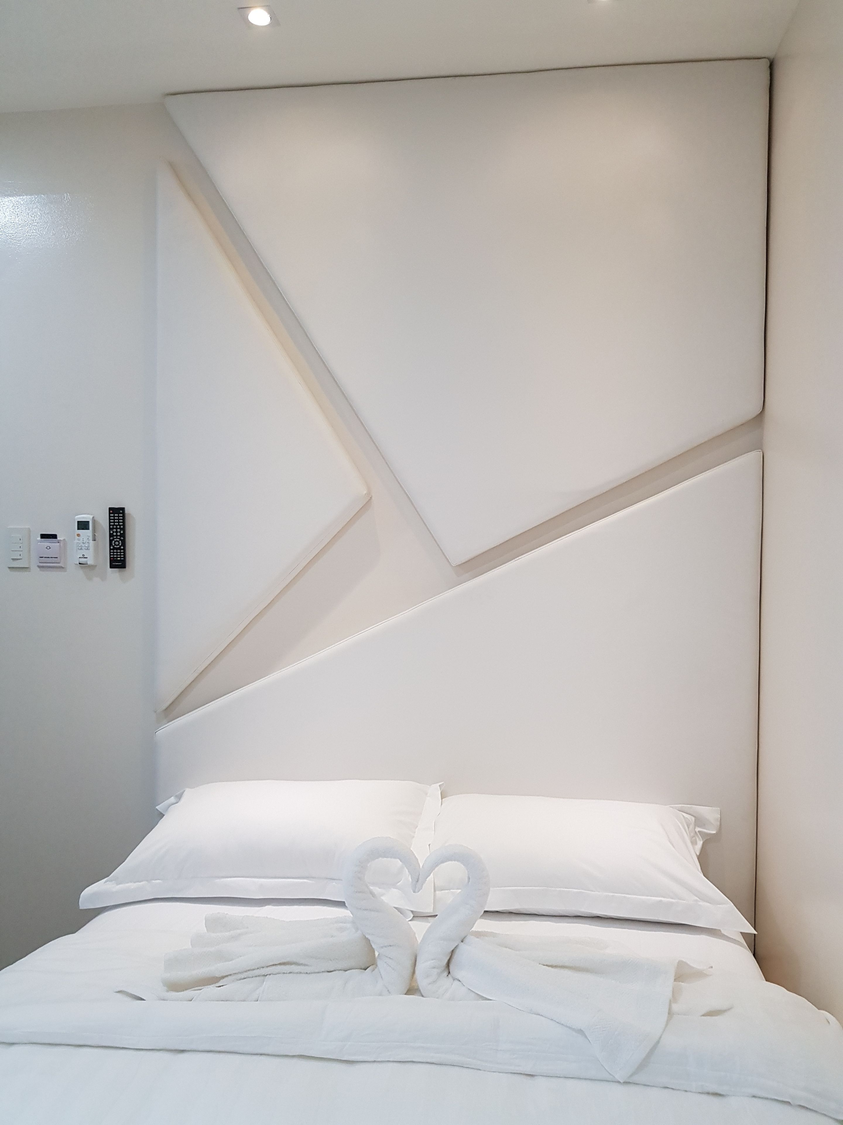 A modern hotel room with a neatly made bed, a heart-shaped towel arrangement, and a geometric wall design.
