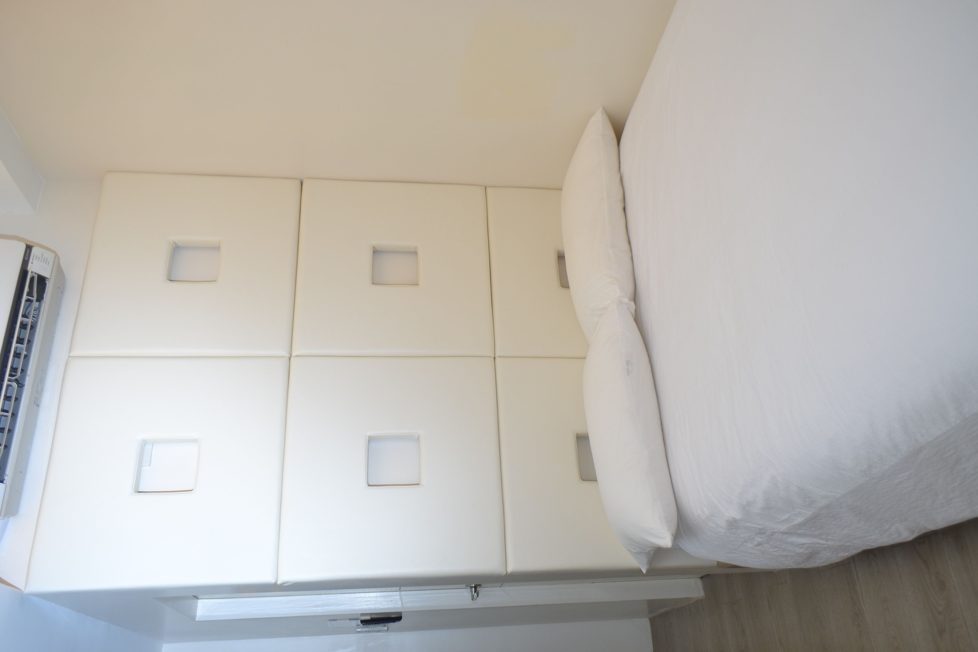 Un lit blanc avec un oreiller, une armoire blanche à quatre portes et une petite fenêtre carrée sur le mur.