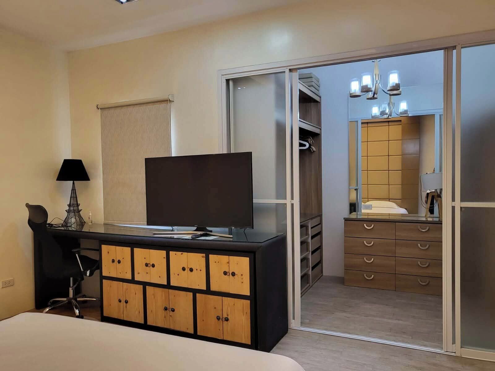 A hotel room with a bed, a TV on a black dresser, a desk with a lamp, and a sliding closet door.
