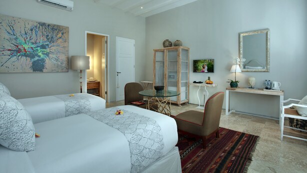 A hotel room with two beds, a desk, a chair, a small table, a cabinet, and a large abstract painting. - Room & Vespa 4 (Seminyak)