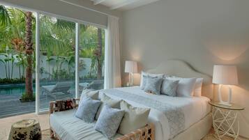 A bedroom with a large bed, two bedside lamps, a wooden headboard, and a view of a pool and palm trees through large windows.
