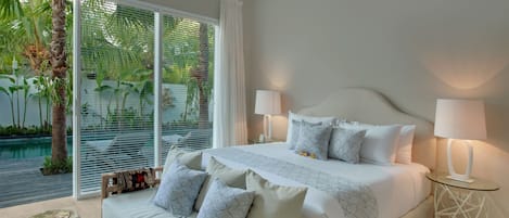 A bedroom with a large bed, two bedside lamps, a wooden headboard, and a view of a pool and palm trees through large windows.