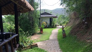A pathway leading to a house with a tiled roof, surrounded by lush greenery and a thatched-roof structure. - Chang Pai (Pai)