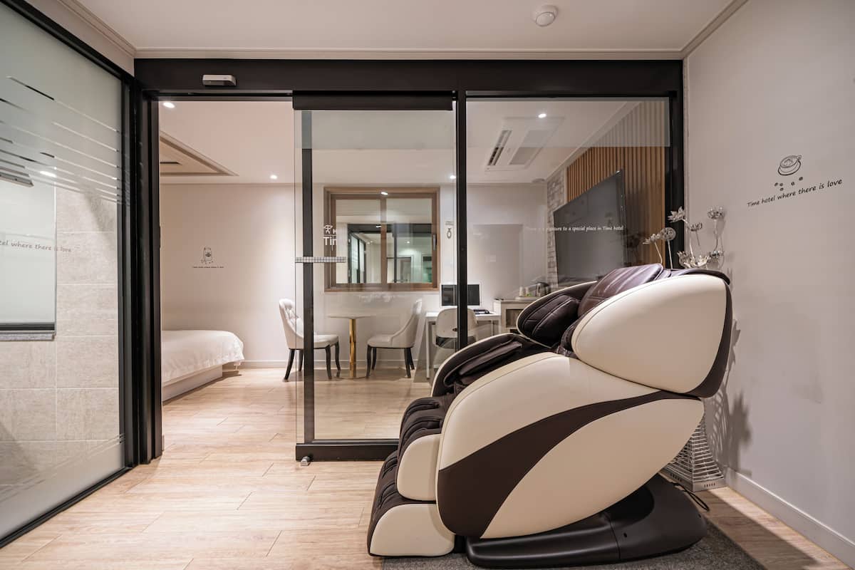 A modern hotel room with a massage chair, a bed, a dining area, and a view through a glass door.