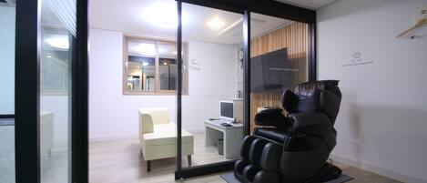 A massage chair in a room with a glass door leading to a seating area and a desk with a computer.