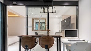 A modern hotel room with a glass wall, a wooden table, two chairs, a desk with a computer, and a decorative vase.