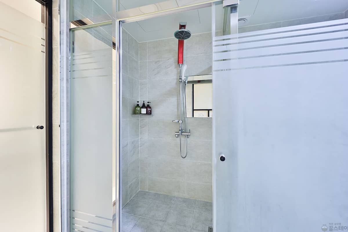 A glass-enclosed shower room with a handheld showerhead, a glass door, and a shelf with toiletries.