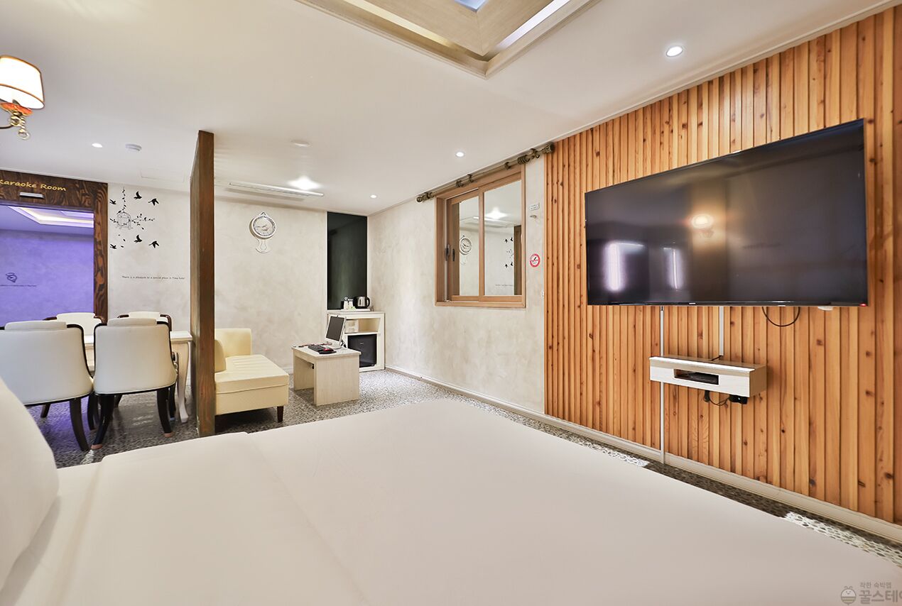 A hotel room with a large bed, a TV mounted on a wooden wall, a small desk with a chair, and a karaoke room sign.