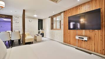 A hotel room with a large bed, a TV mounted on a wooden wall, a small desk with a chair, and a karaoke room sign.