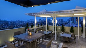 A rooftop terrace with tables and chairs, a bottle of wine, and a candle, overlooking a cityscape at dusk.
