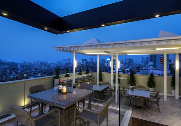 Une terrasse sur le toit, avec des tables et des chaises, une bouteille de vin et une bougie, d’où l’on peut admirer la vue sur la ville au crépuscule.