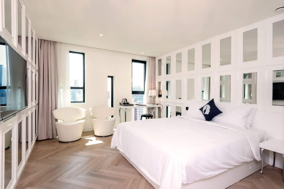 Una habitación de hotel moderna con una cama grande, una zona de estar con sillas, una mesita y ventanas amplias.