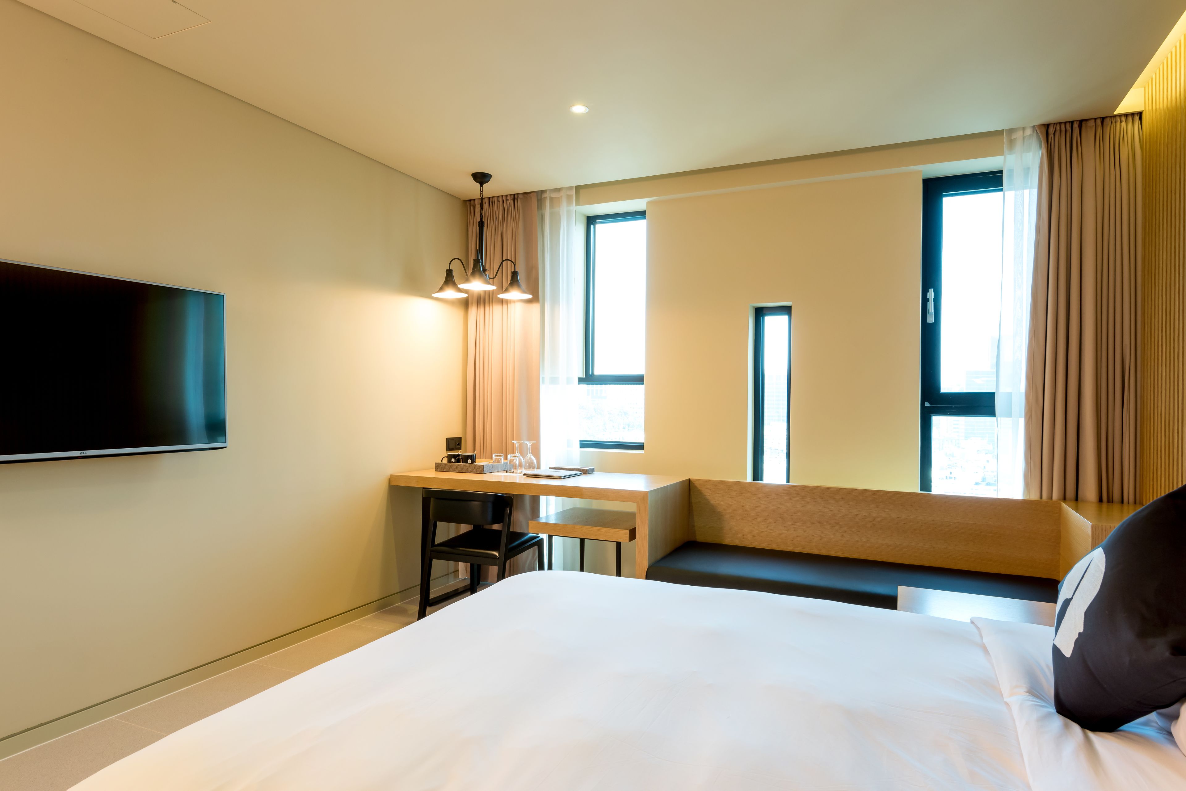 A hotel room with a large bed, a desk with a chair, a flat-screen TV mounted on the wall, and a window with curtains.