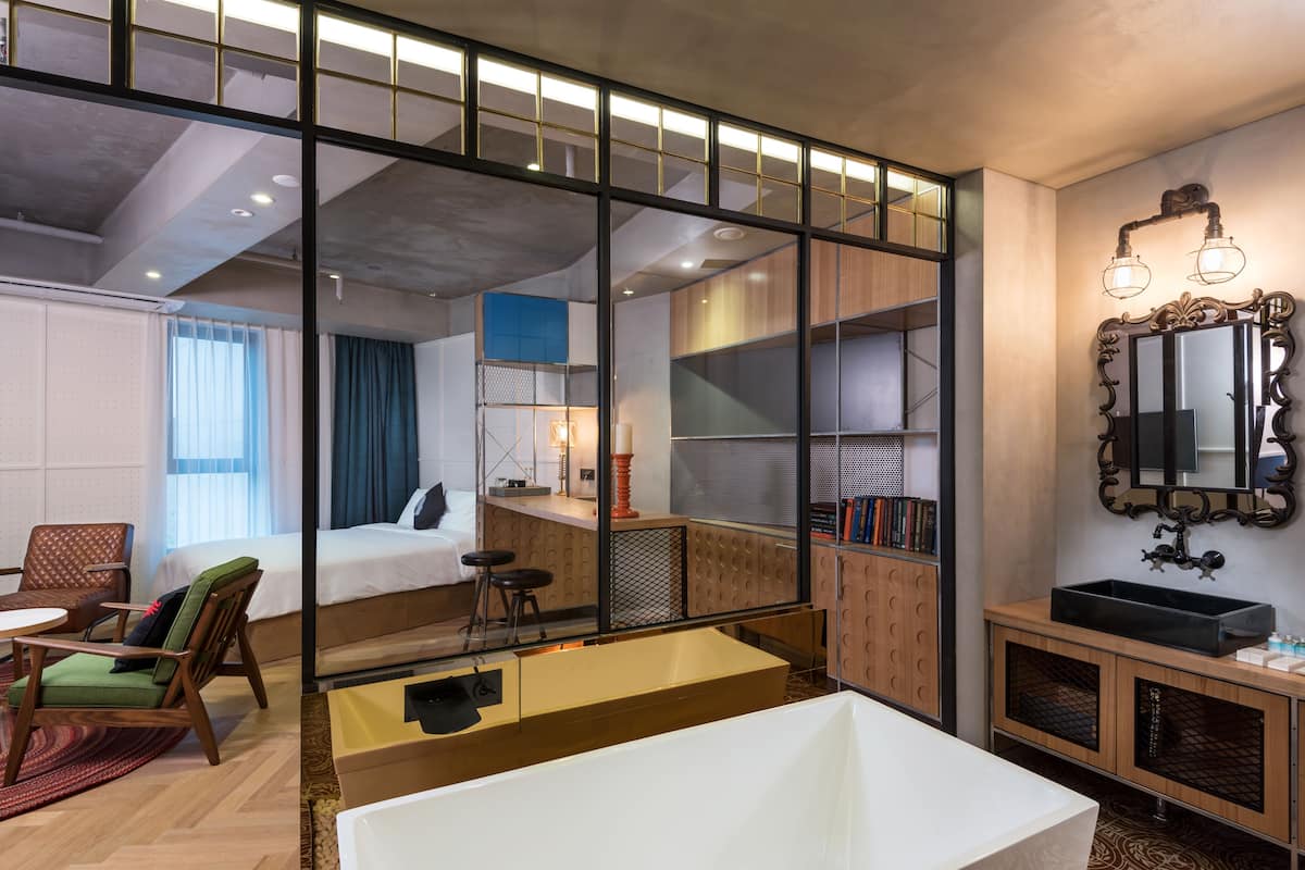 Una habitación de hotel moderna con una cama grande, una zona de estar con mesa y sillas, una pequeña cocina y un baño con un espejo grande y lavamanos.