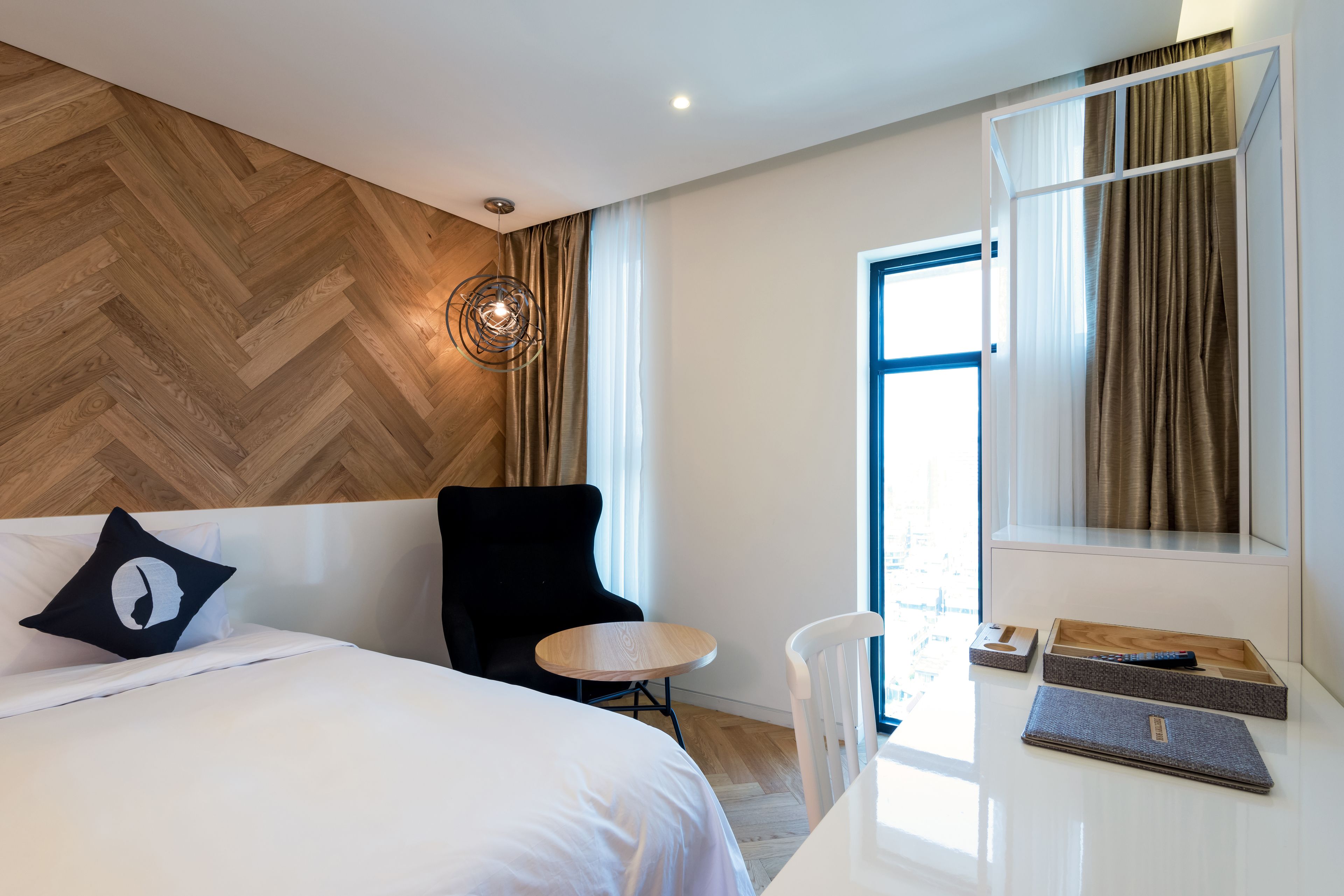 A modern hotel room with a large bed, a small round table, a chair, and a desk with a computer and a box.