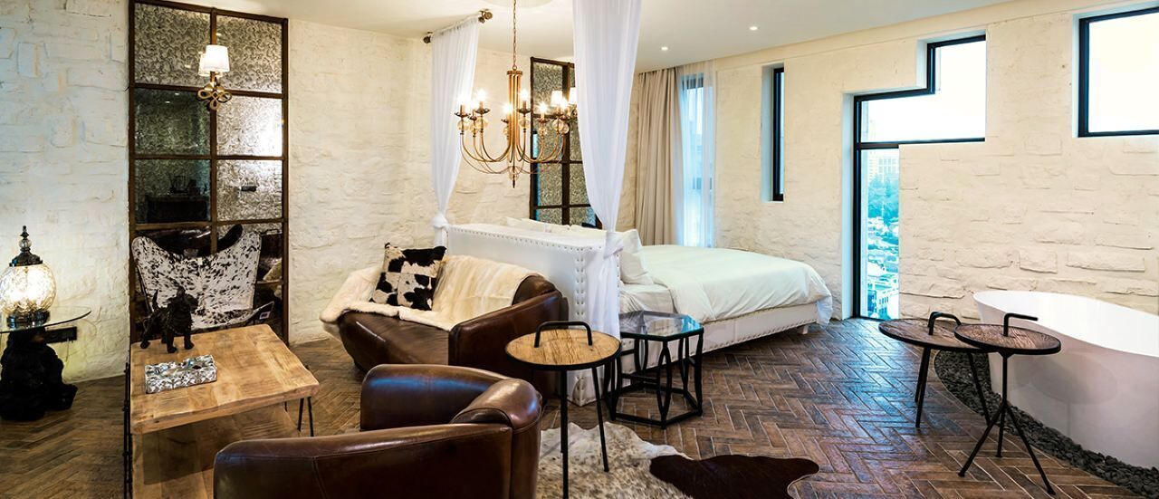 A bedroom with a bed, a sitting area with a table and chairs, a large window, and a view of the city.