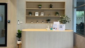 Un mostrador de recepción con acabado en madera clara, tres lámparas colgantes y estantes con plantas y cuadros enmarcados.