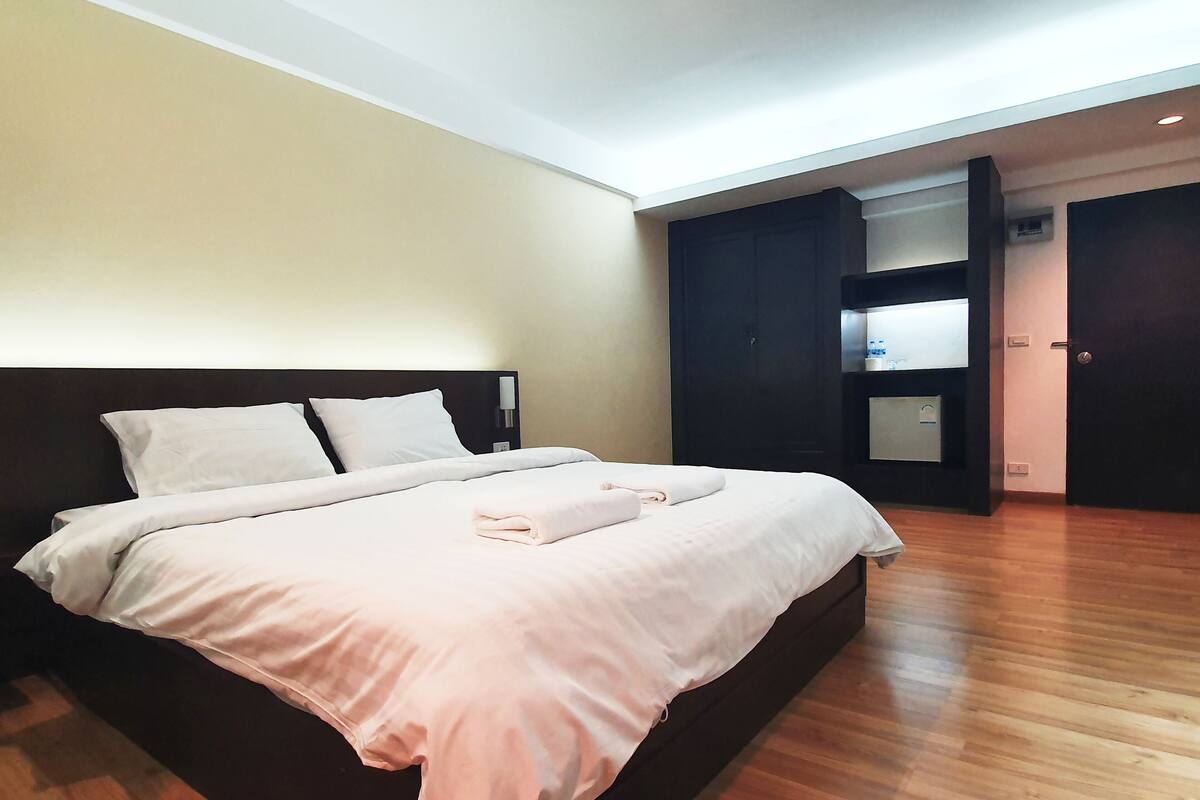 A neatly made bed with pillows and a folded towel, a wooden floor, and a dark-colored wardrobe with a mirror and a small shelf.