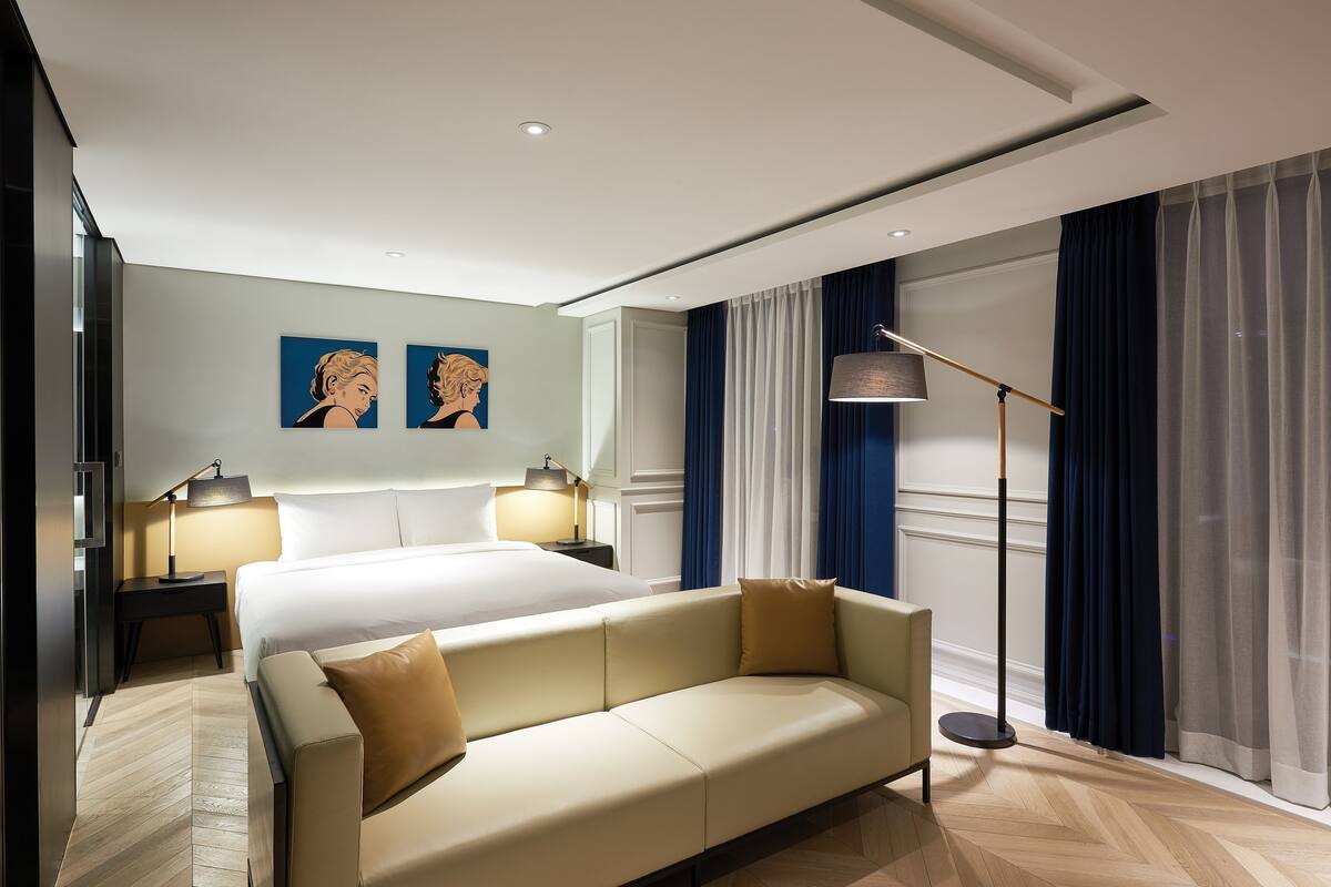 A modern hotel room with a large bed, a sofa, two bedside tables with lamps, and artwork on the wall.
