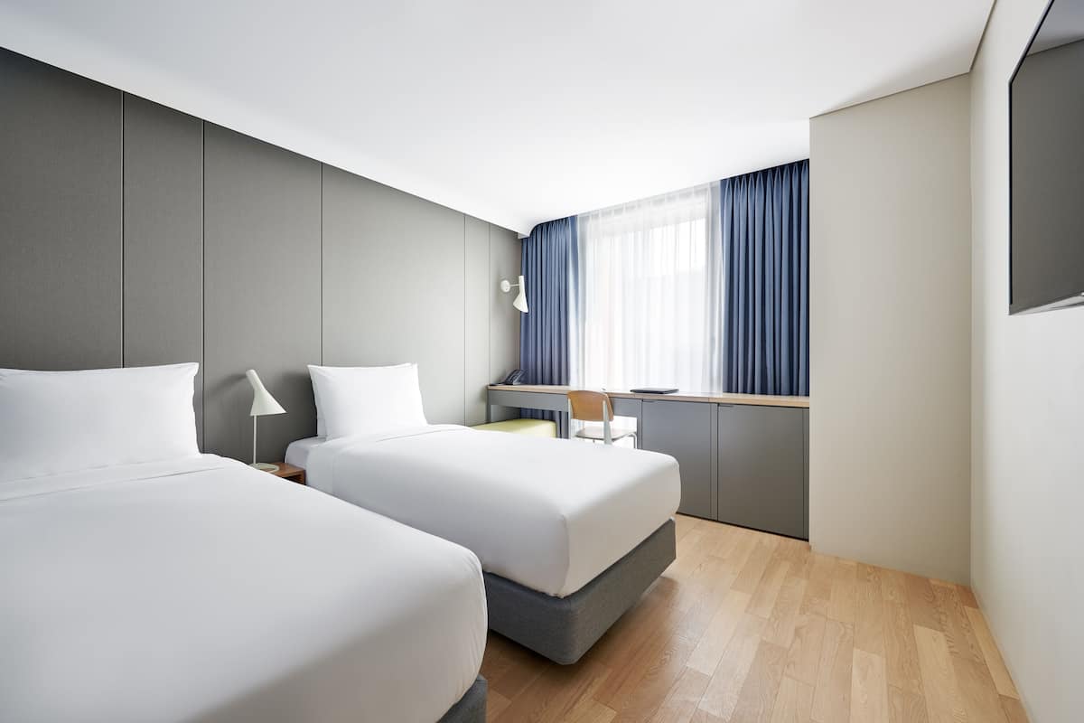A modern hotel room with two beds, a desk, and a large window with curtains.