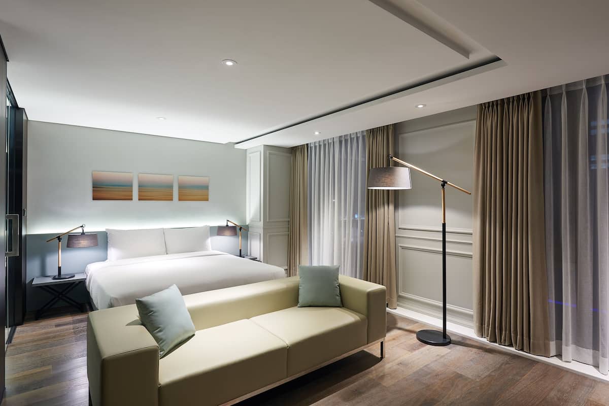 A modern hotel room with a large bed, a sofa, a standing lamp, and a view of the city through curtains.