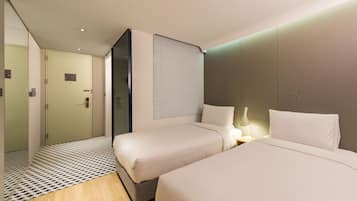 A modern hotel room with two beds, a bedside table with a lamp, a large window with blinds, and a door leading to a bathroom.